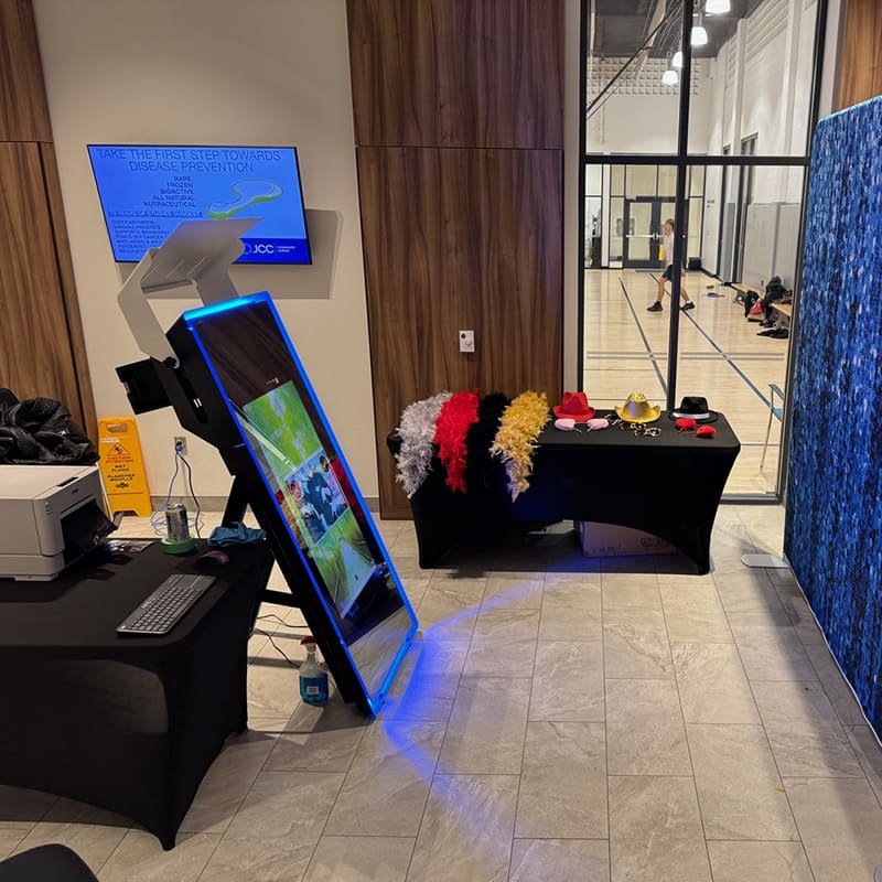 Photo booth setup with touchscreen mirror, props table, and blue sequin backdrop in an event space.