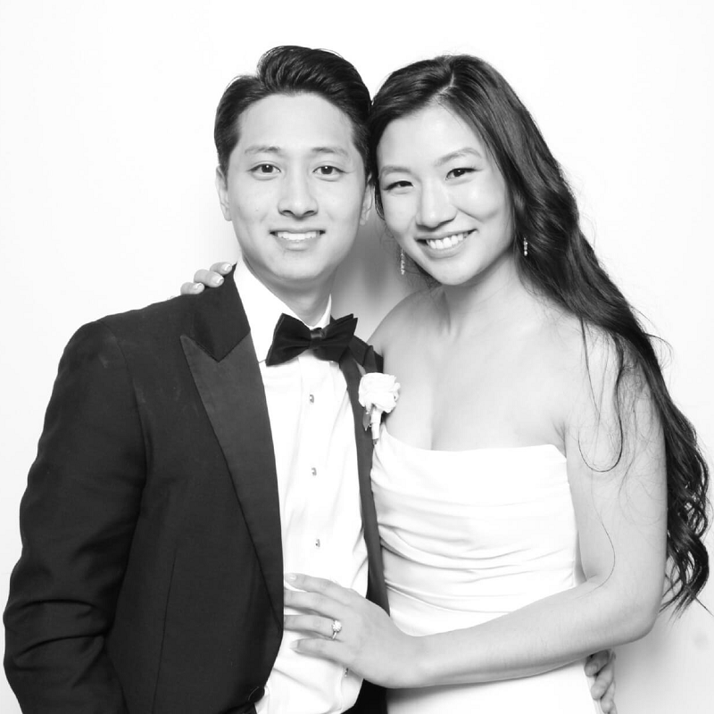 Black and white photo booth portrait of a smiling couple in formal wedding attire, posing closely together against a white backdrop.