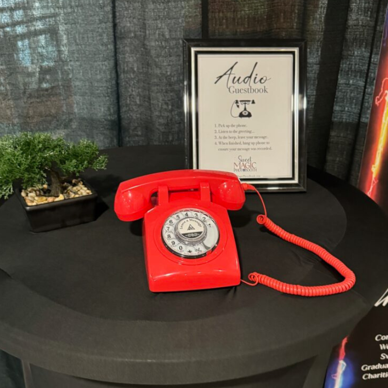 Bright red rotary phone audio guestbook displayed on a black table with framed instructions.