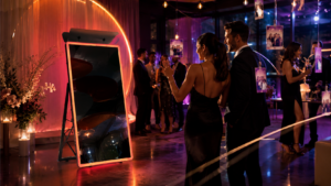 Group of guests in formal attire mingling at a chic party beside a glowing neon-framed photo booth and hanging photos.